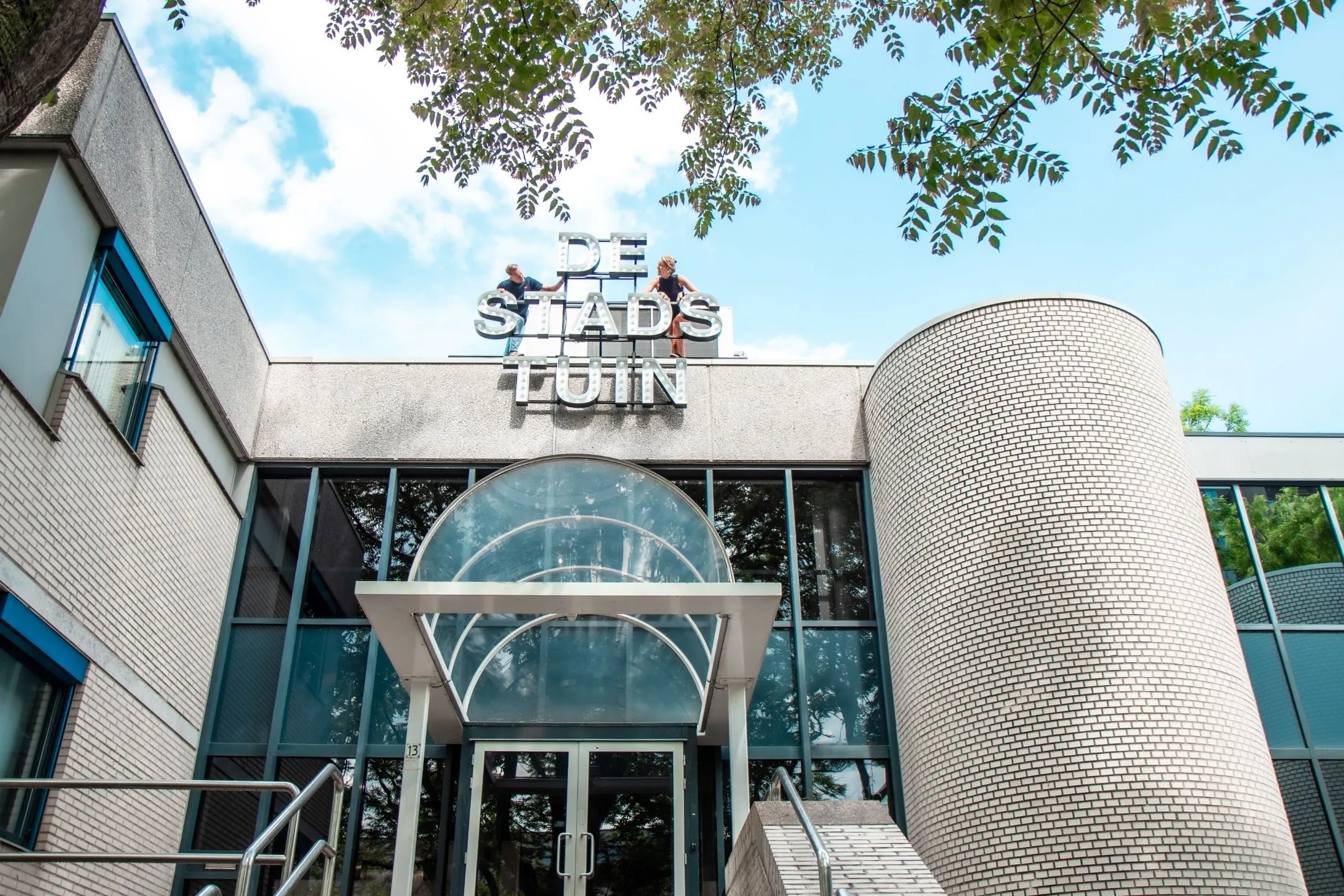 Vooraanzicht van het gebouw aan de Bernadottelaan 10 met de ingang van De Stadstuin en twee personen op het dak bij het gevelbord.