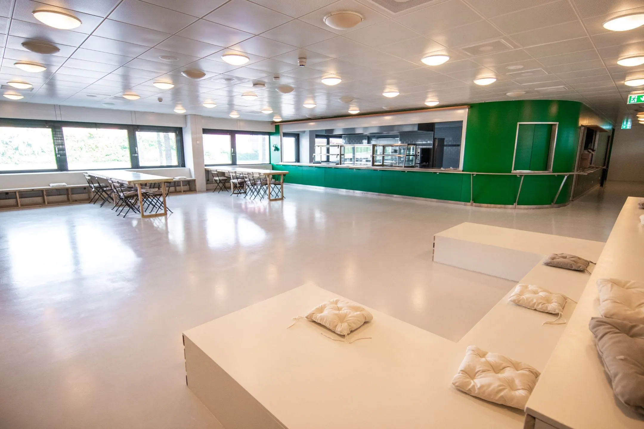 Moderne kantineruimte aan de Bernadottelaan met groene uitgiftebalie, tafels, stoelen en zitkussens op een witte tribune.