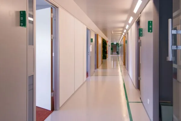 Heldere gang in het gebouw aan de Bernadottelaan 7 met openstaande kantoordeuren en groene kamernummers aan de muren.