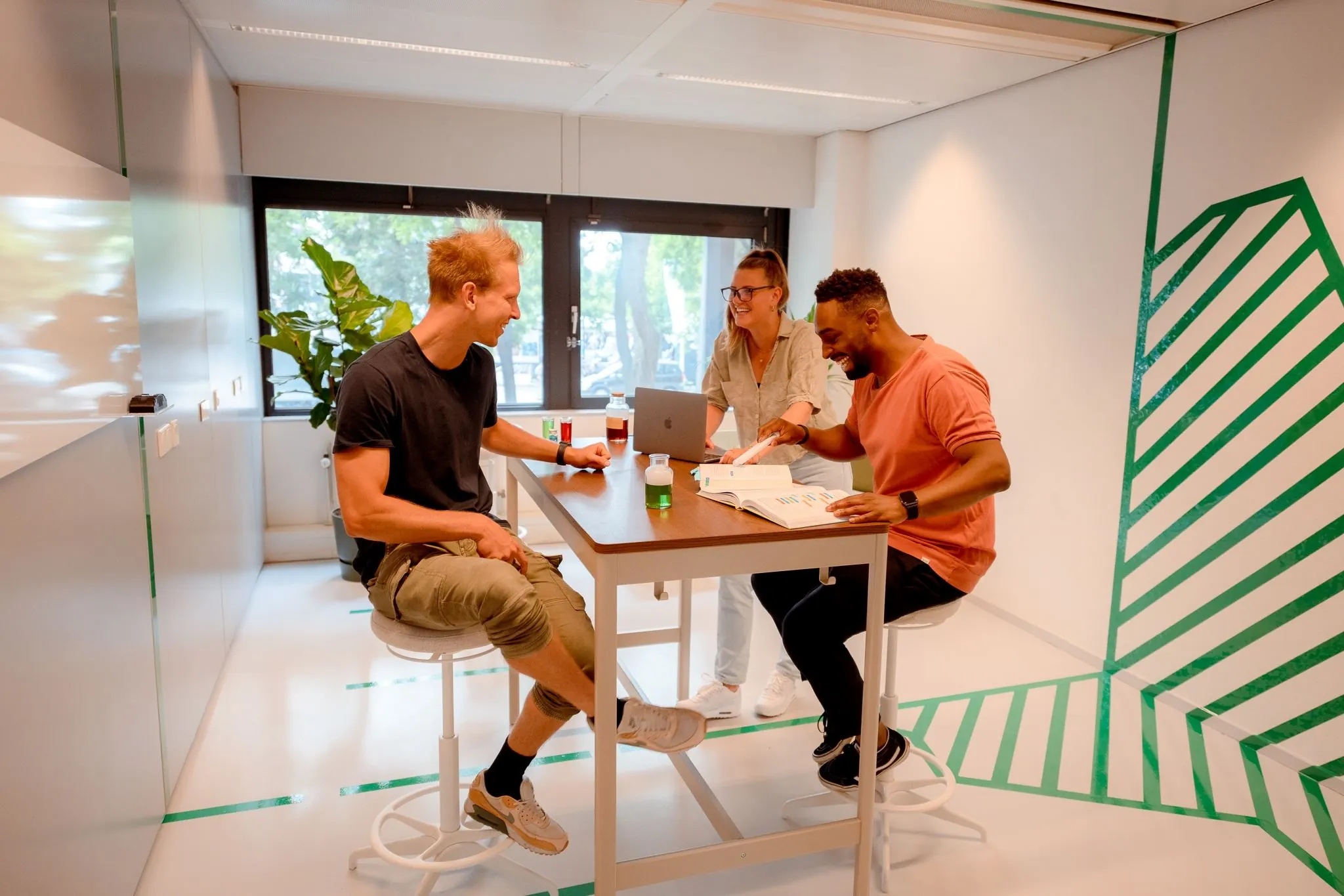 Drie mensen werken en lachen samen aan een hoge tafel in een moderne kantoorruimte aan de Bernadottelaan.