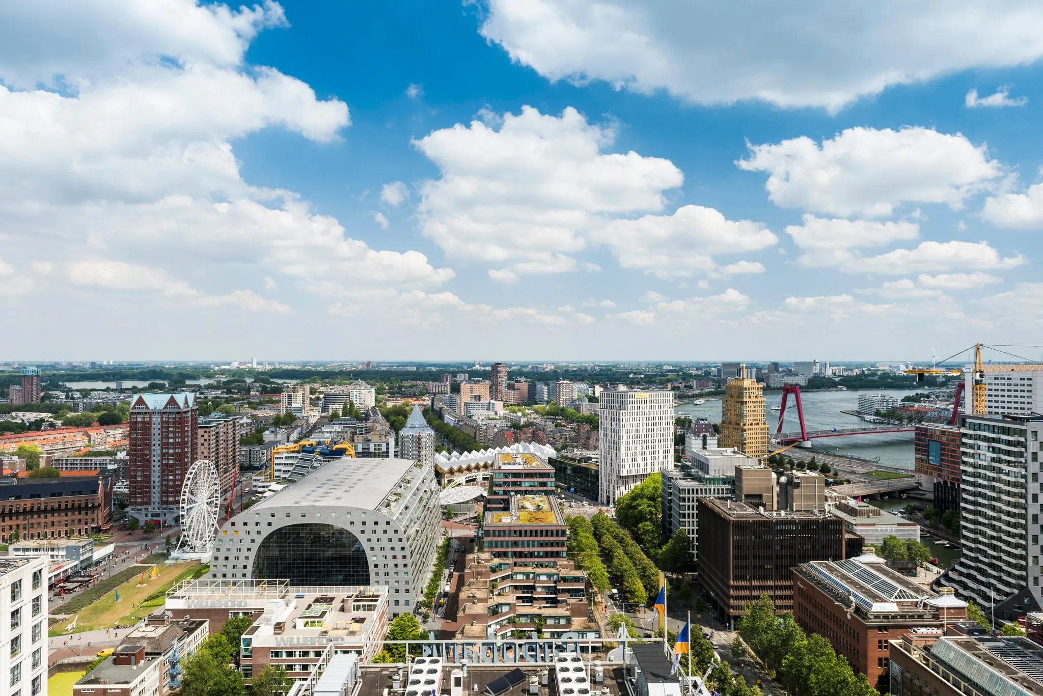 Luchtfoto van Rotterdam met de Markthal, Kubuswoningen en de Willemsbrug zichtbaar onder een deels bewolkte blauwe lucht.