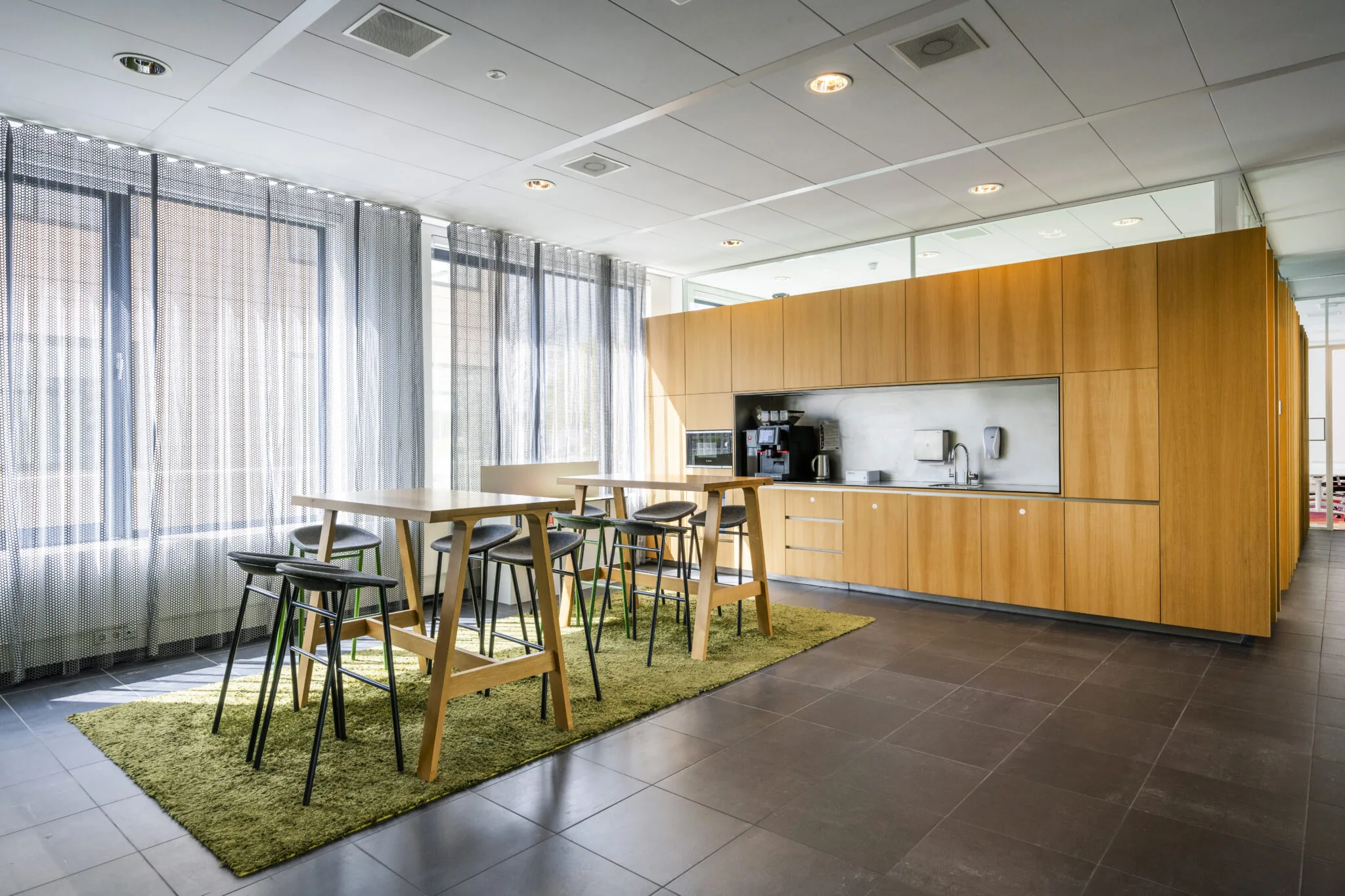 Moderne kantoorkeuken aan de Boeingavenue met houten kasten, hoge tafels, barkrukken en een koffieapparaat.