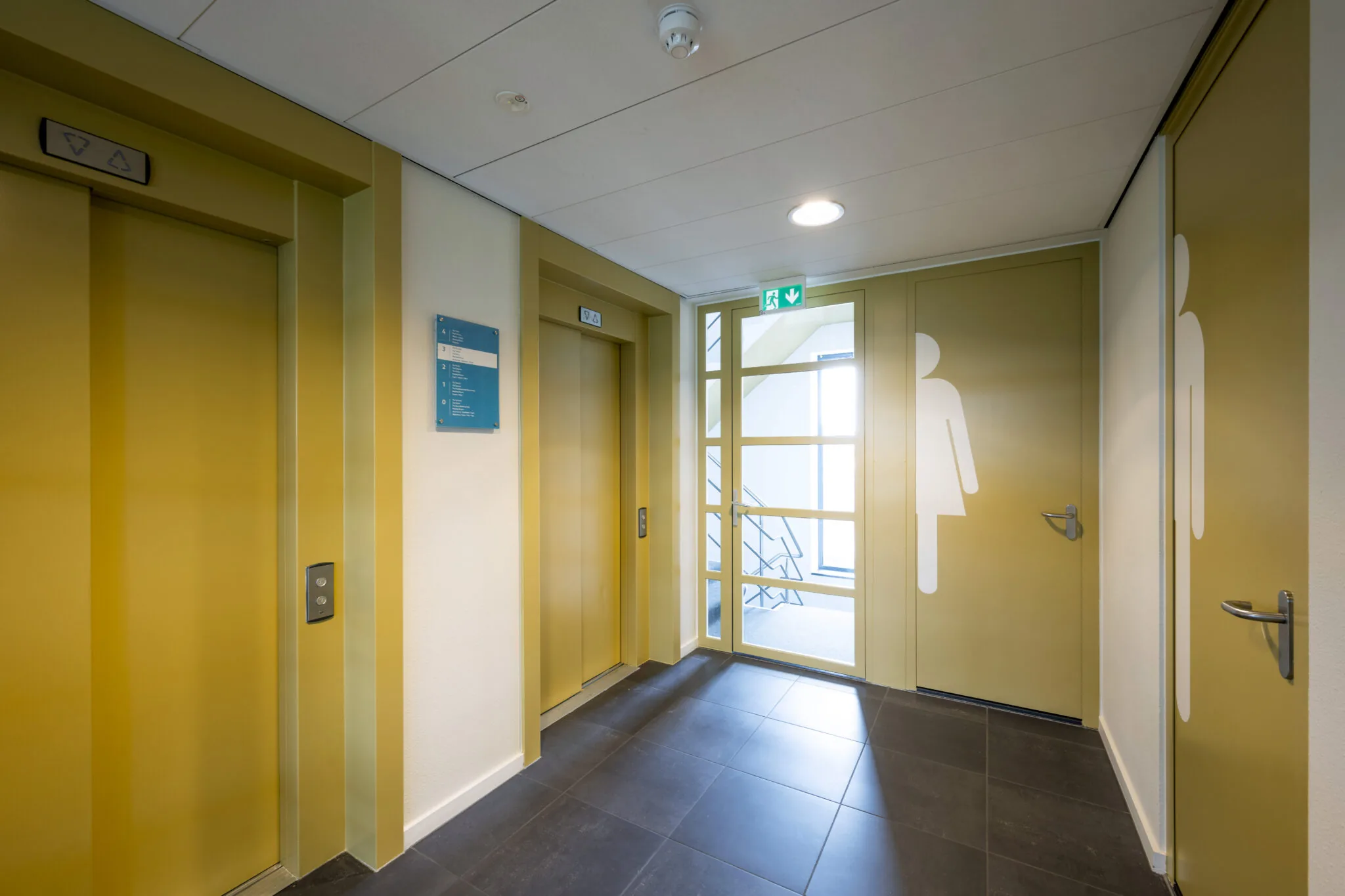 Gele liftlobby in een kantoorpand aan de Boeingavenue met twee liften, toiletten en een trapdeur.
