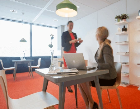 Twee zakenvrouwen overleggen in een moderne kantoorruimte met feloranje vloerbedekking.