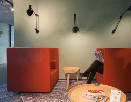 Moderne loungehoek met oranje zitbanken, een houten tafeltje en een persoon die een tijdschrift leest.