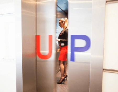 Vrouw in een rode rok stapt een lift binnen met de letters "UP" op de deuren.