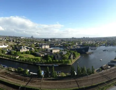 Panoramisch uitzicht op een stad met een rivier, groene zones, spoorlijnen en moderne gebouwen onder een blauwe lucht.