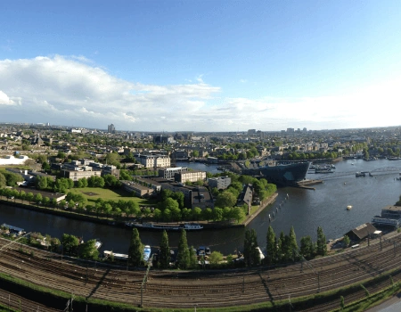 Panoramisch uitzicht op een stad met een rivier, groene zones, spoorlijnen en moderne gebouwen onder een blauwe lucht.