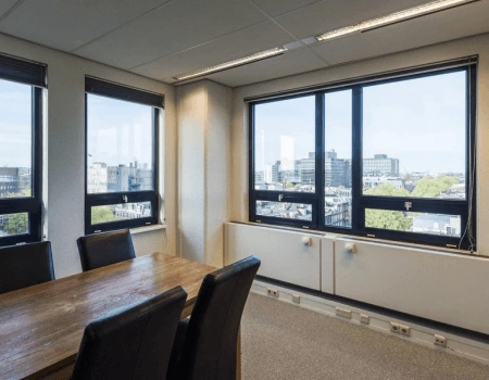Vergaderruimte met houten tafel en zwarte stoelen, met uitzicht op een stedelijk landschap door grote ramen.
