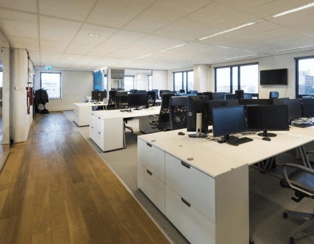 Moderne, open kantoortuin met bureaus, computers en grote ramen met uitzicht op de stad.