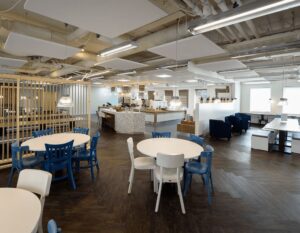 Moderne en ruime bedrijfsrestaurant met ronde tafels, blauwe en witte stoelen, en een open keuken in het midden.