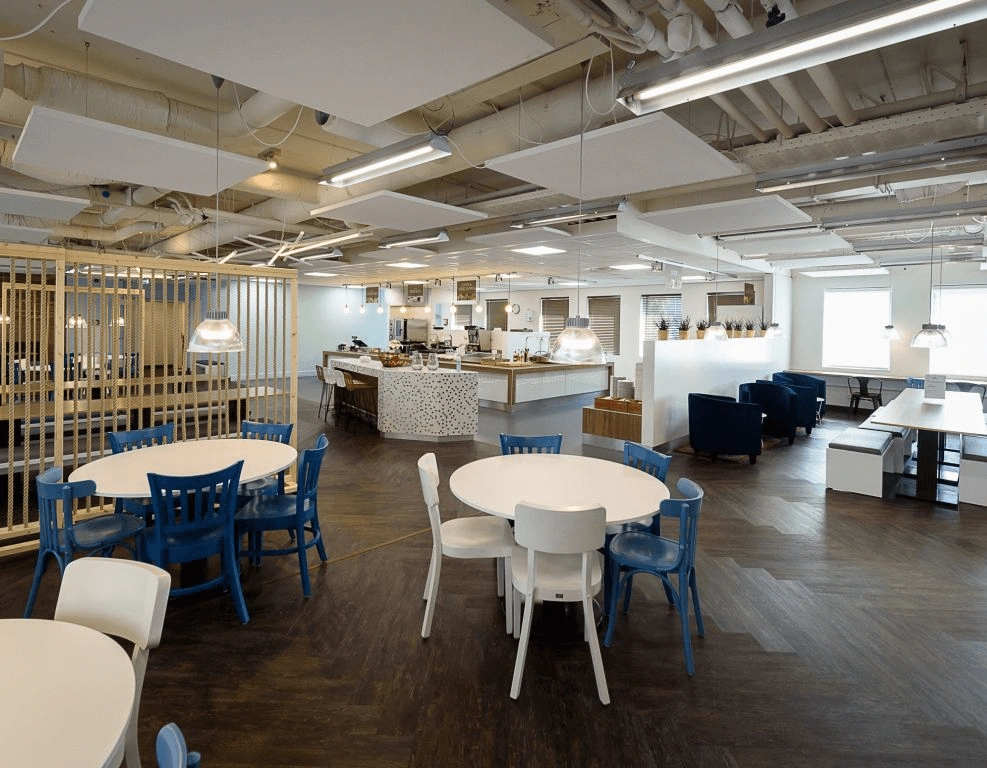 Moderne en ruime bedrijfsrestaurant met ronde tafels, blauwe en witte stoelen, en een open keuken in het midden.