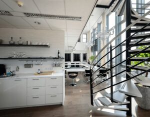 Moderne kantoorruimte met een open keuken, design trap en grote ramen die veel daglicht binnenlaten.
