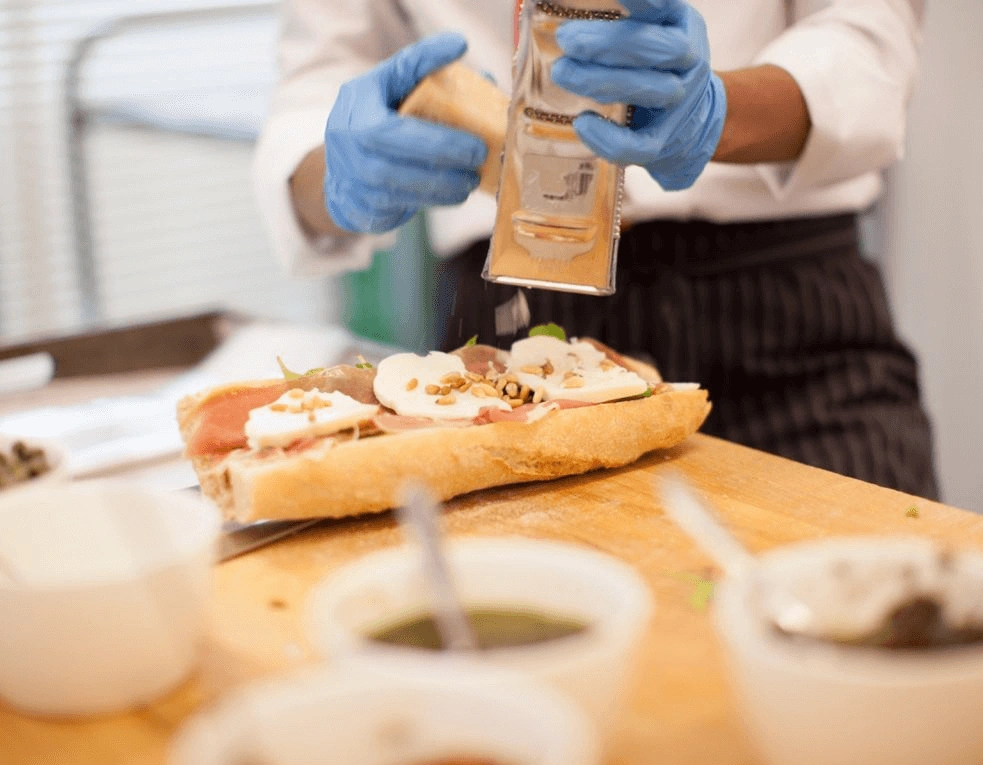 Kok met blauwe handschoenen raspt kaas over een belegde baguette op een houten werkblad.