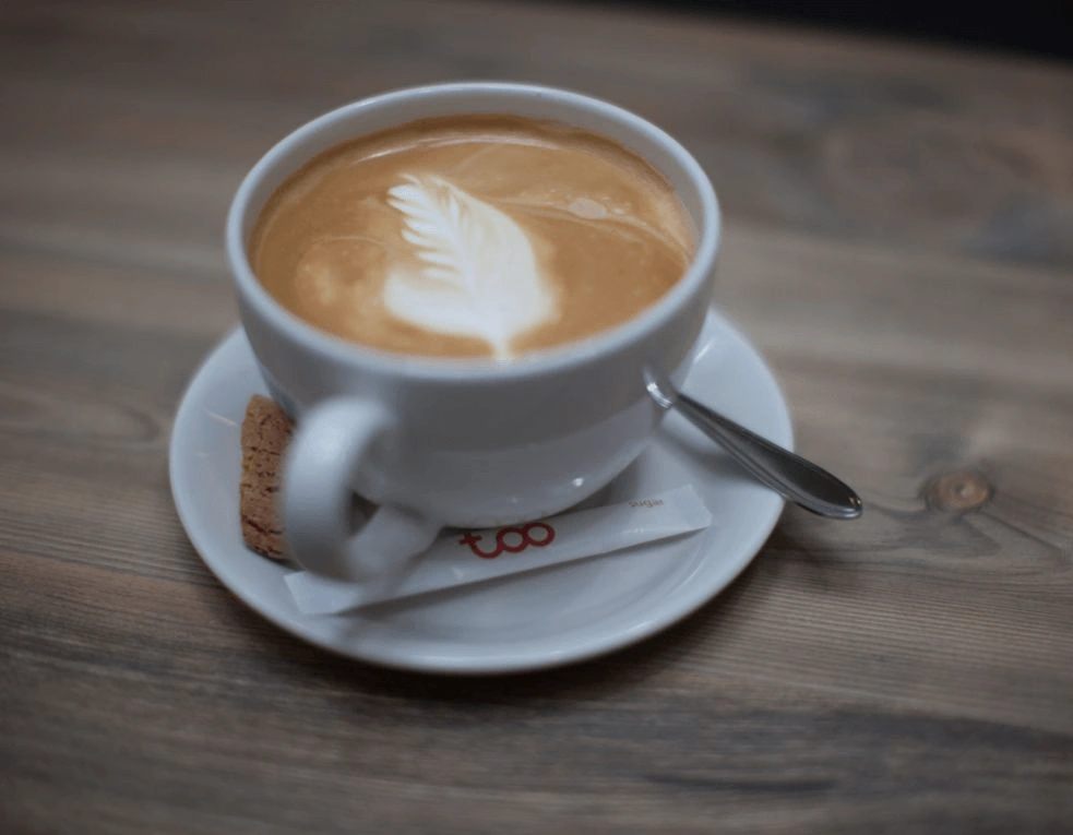 Een cappuccino met latte art in de vorm van een blad, geserveerd op een schoteltje met een lepel, suikerstick en biscuitje.