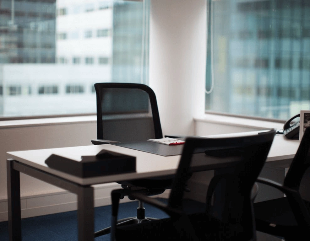 Moderne, minimalistische kantoorruimte met bureaus, bureaustoelen en grote ramen met uitzicht op kantoorgebouwen.