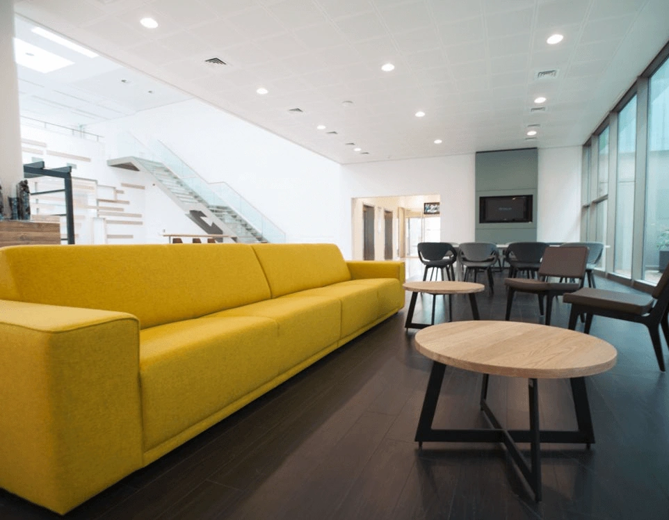 Moderne wachtruimte met een lange gele bank, houten salontafels en zwarte stoelen op een donkere houten vloer.