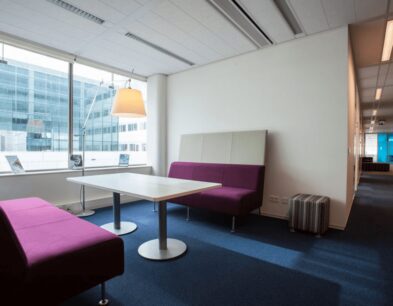Moderne kantoorlounge met paarse banken, een witte tafel en grote ramen met uitzicht op een kantoorgebouw.