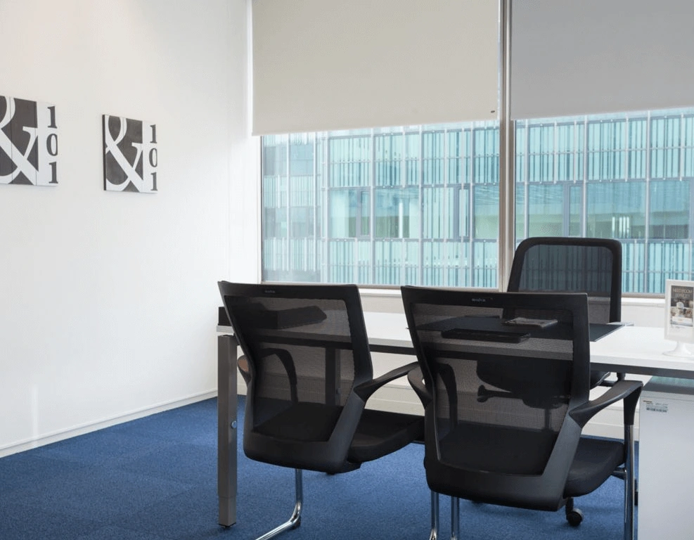 Modern kantoor met drie bureaustoelen, een bureau, wanddecoratie en uitzicht op een glazen gevel van een naastgelegen gebouw.