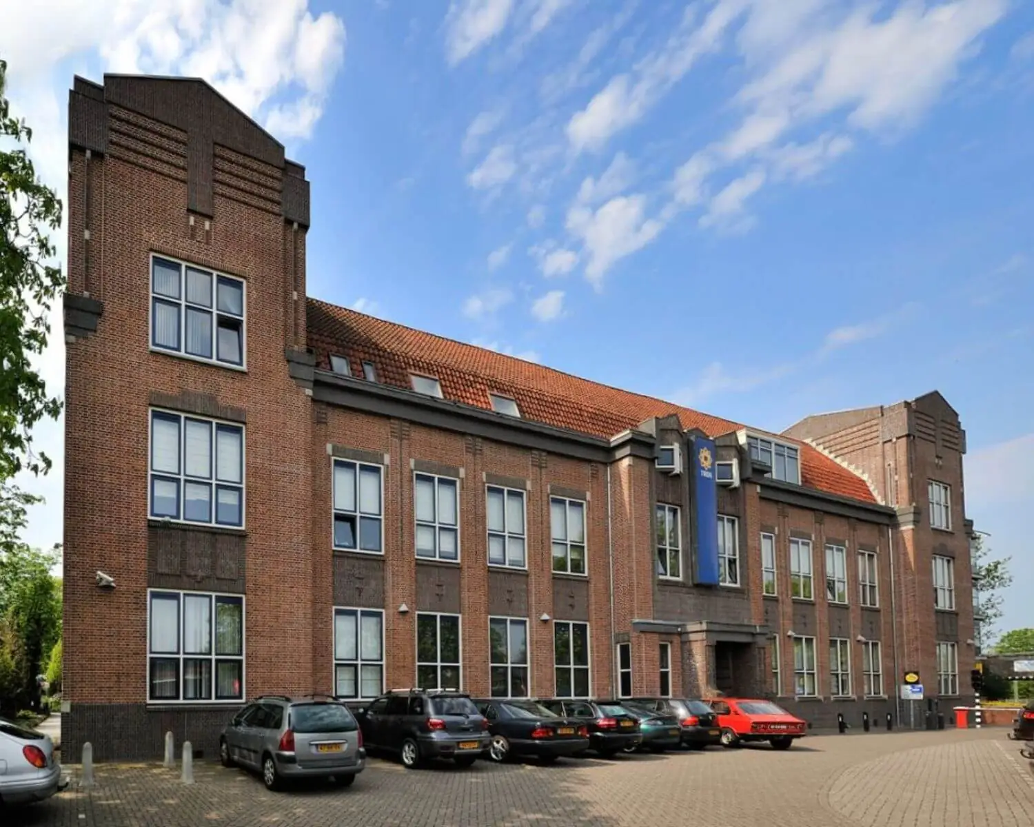 Bakstenen kantoorgebouw met een rood pannendak en een politie-embleem aan de gevel, gezien vanaf de parkeerplaats.