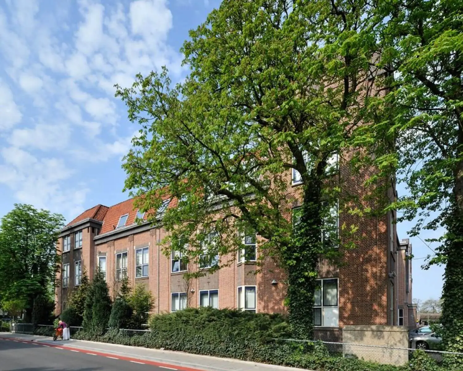 Bakstenen schoolgebouw met rode dakpannen, omgeven door groene bomen en struiken op een zonnige dag.