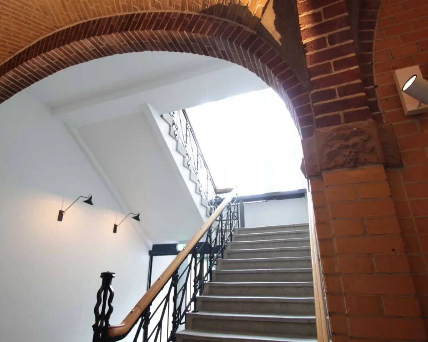 Trapopgang met een metalen leuning en bakstenen boog in een gebouw met veel natuurlijk licht.