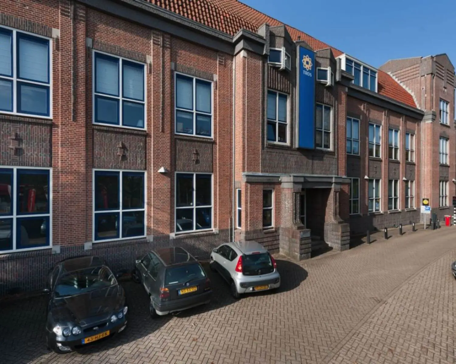 Bakstenen kantoorgebouw met TROS-logo aan de gevel en geparkeerde auto’s op de voorgrond.