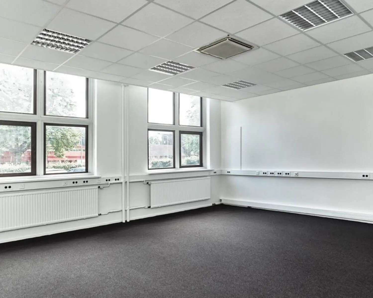 Leeg, licht klaslokaal met grote ramen, witte muren en donker tapijt.