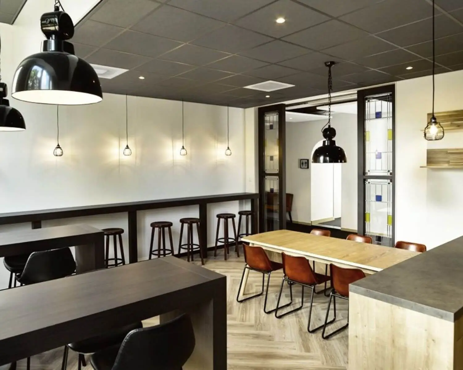 Modern ingerichte kantineruimte met houten tafels, zwarte stoelen en industriële hanglampen.