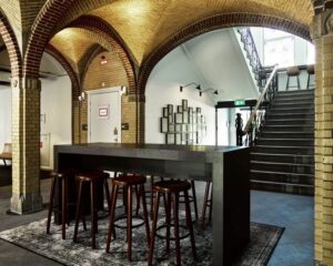 Modern ingerichte ruimte met hoge tafel en barkrukken onder bakstenen gewelven naast een trap.