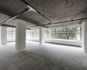 Lege, industriële kantoorruimte met betonnen plafond, grote ramen en witte pilaren.