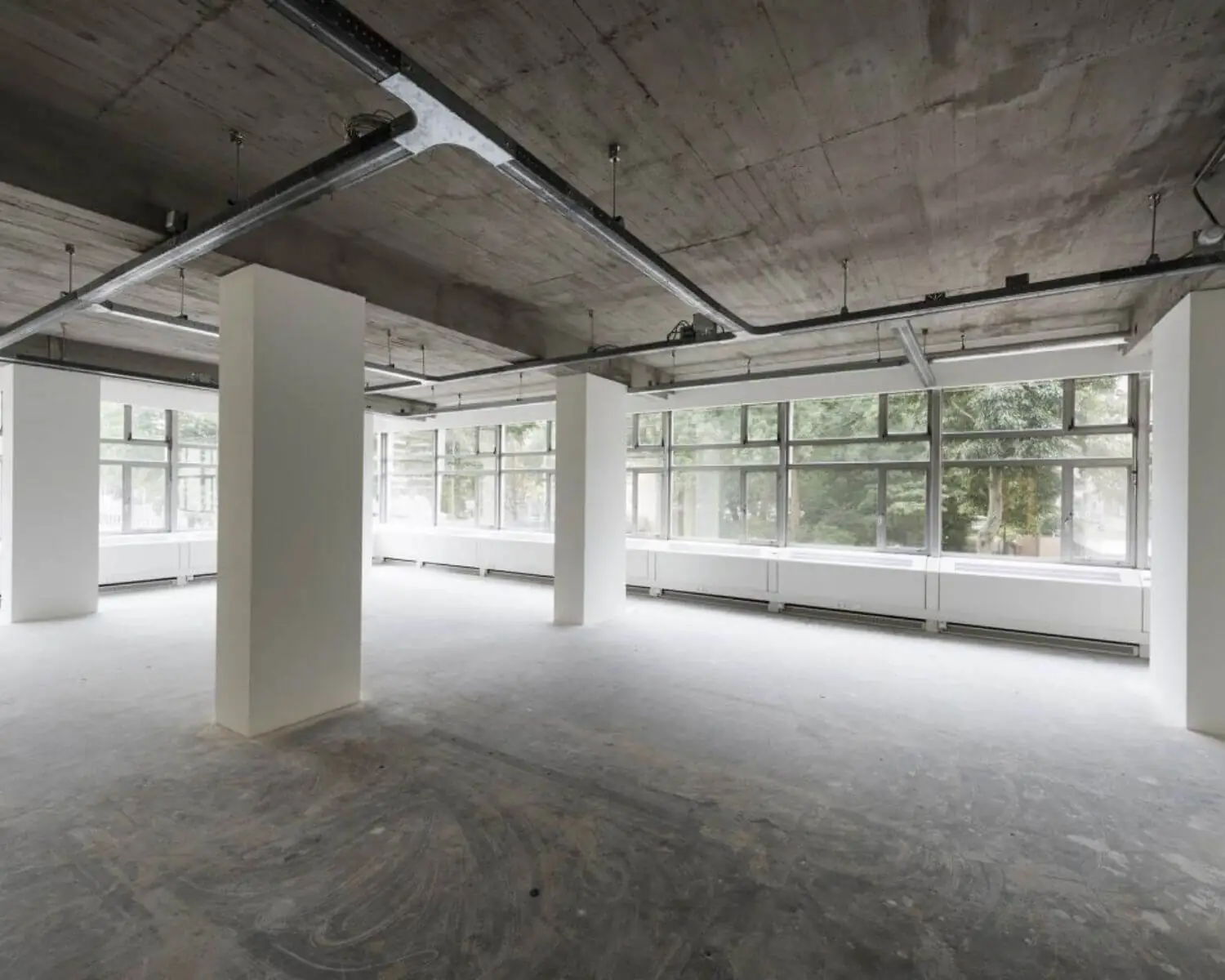 Lege, industriële kantoorruimte met betonnen plafond, grote ramen en witte pilaren.