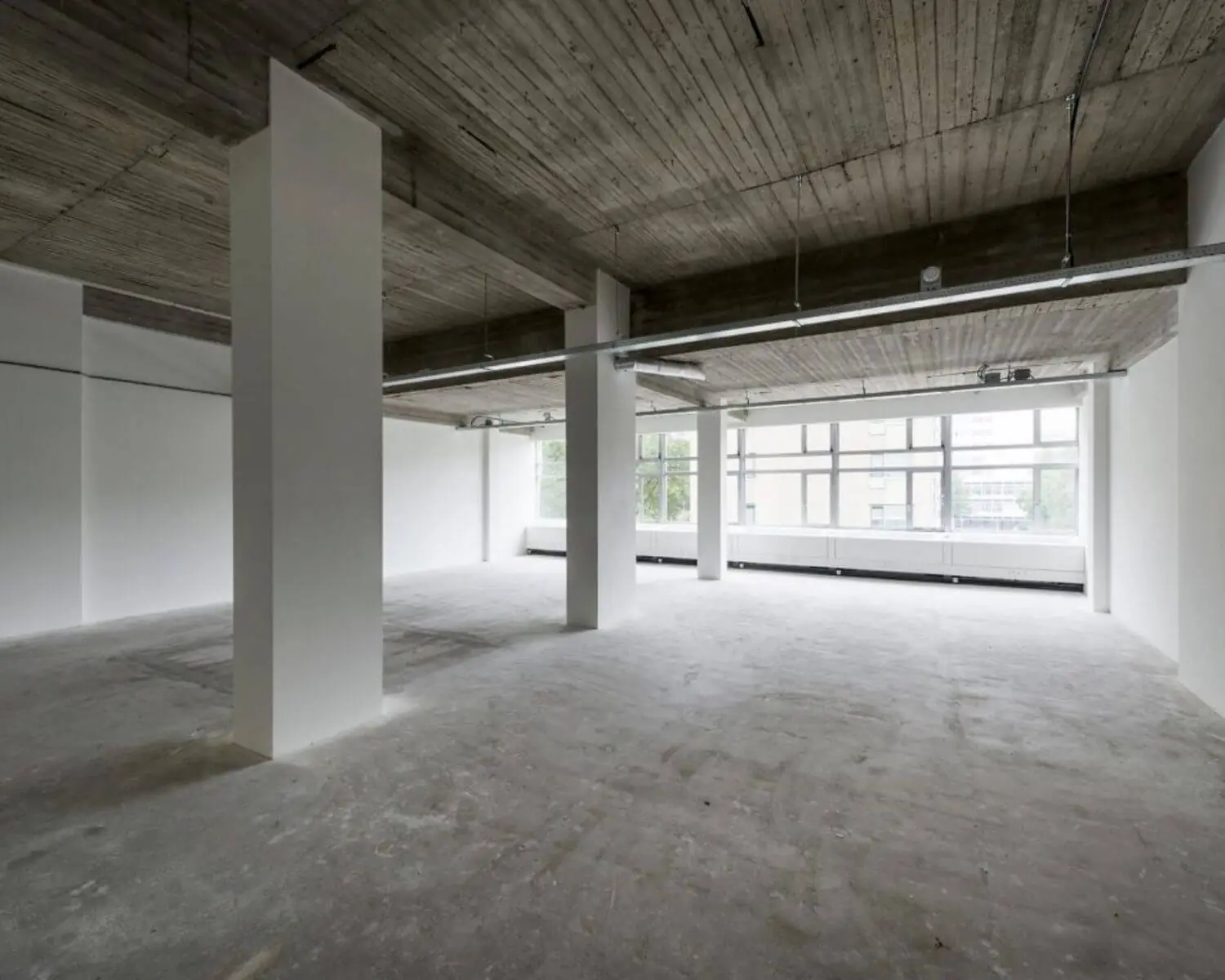 Leeg industrieel kantoor met betonnen vloer, houten plafond en grote ramen met uitzicht op buiten.