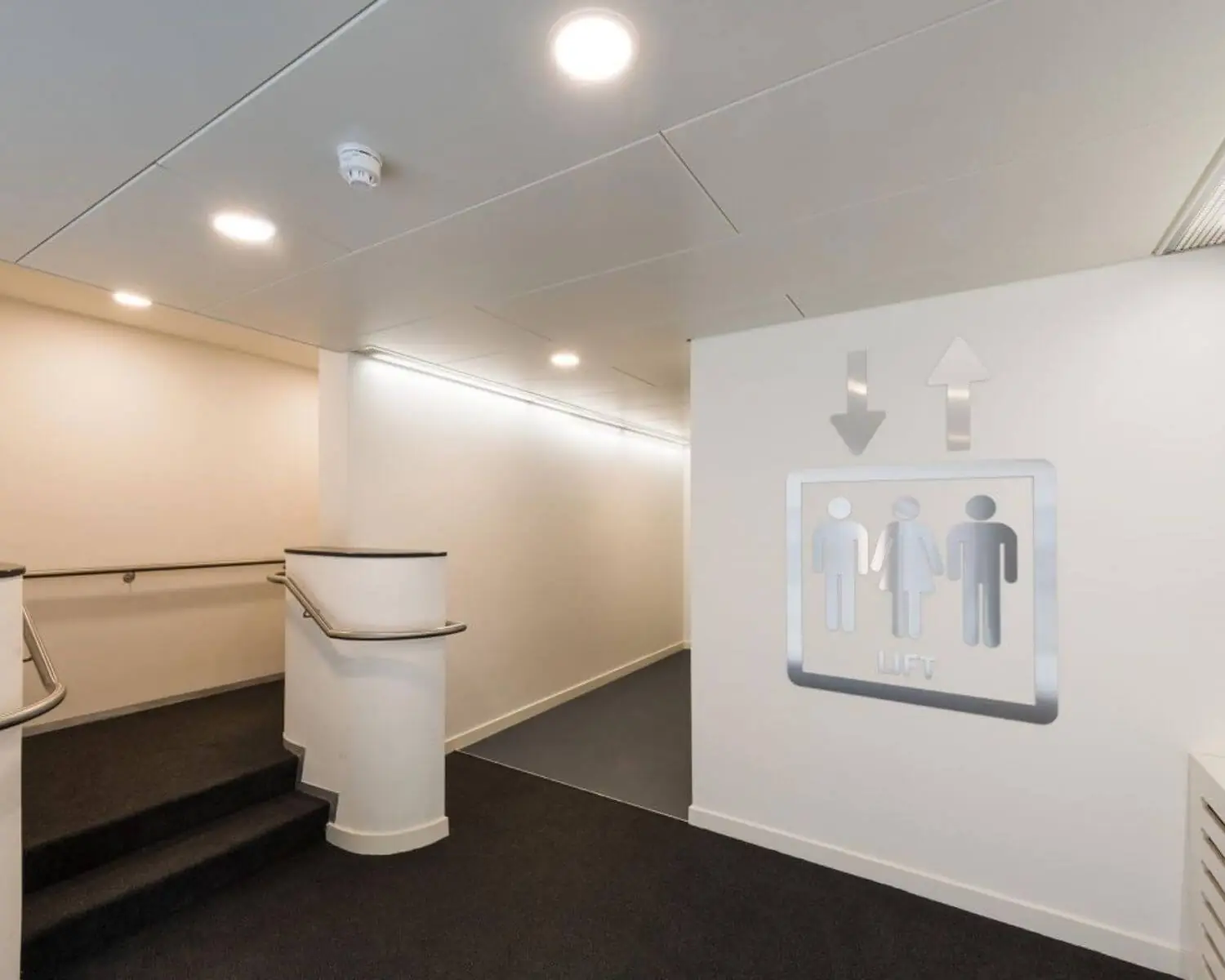 Verlichte gang met trap en helling, met een pictogram dat de richting naar de lift aangeeft.