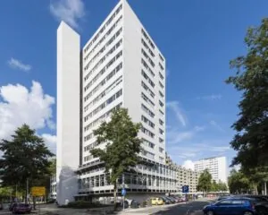 Wit hoog appartementencomplex op een hoek, omringd door bomen en geparkeerde auto's onder een blauwe lucht.