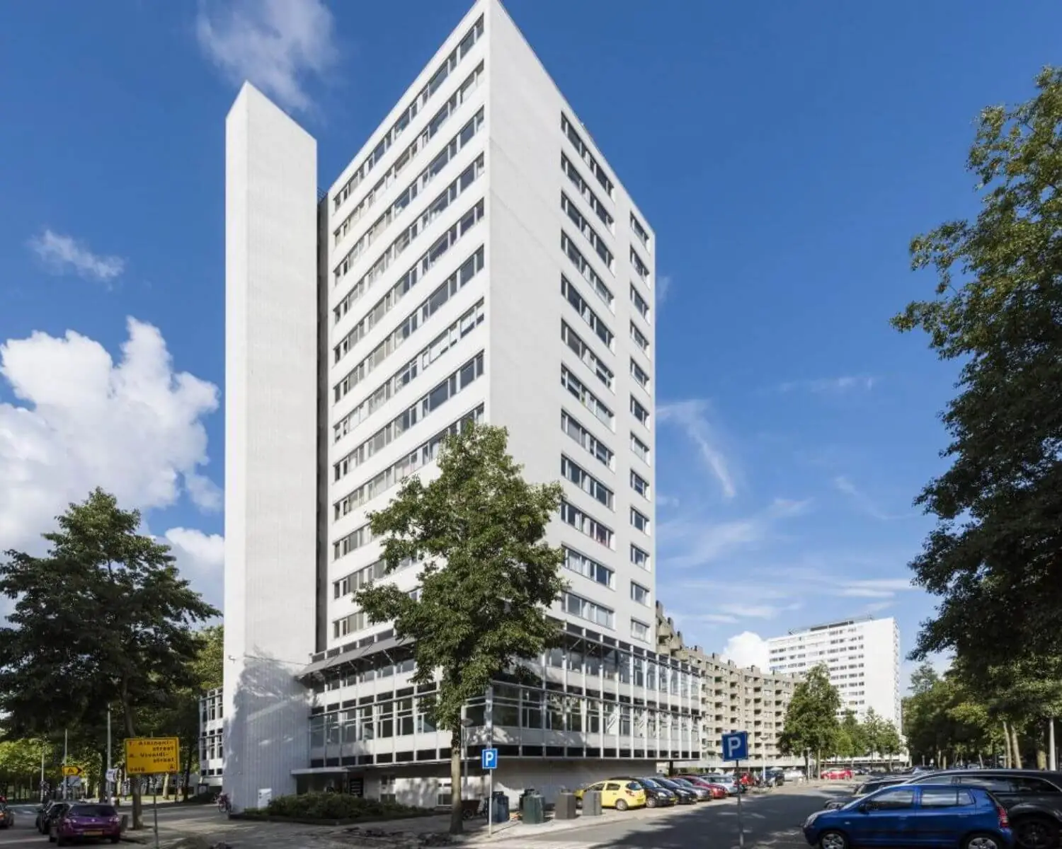 Wit hoog appartementencomplex op een hoek, omringd door bomen en geparkeerde auto's onder een blauwe lucht.