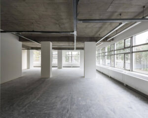 Lege kantoorruimte met betonnen plafond, steunpilaren en grote ramen langs de zijkant.
