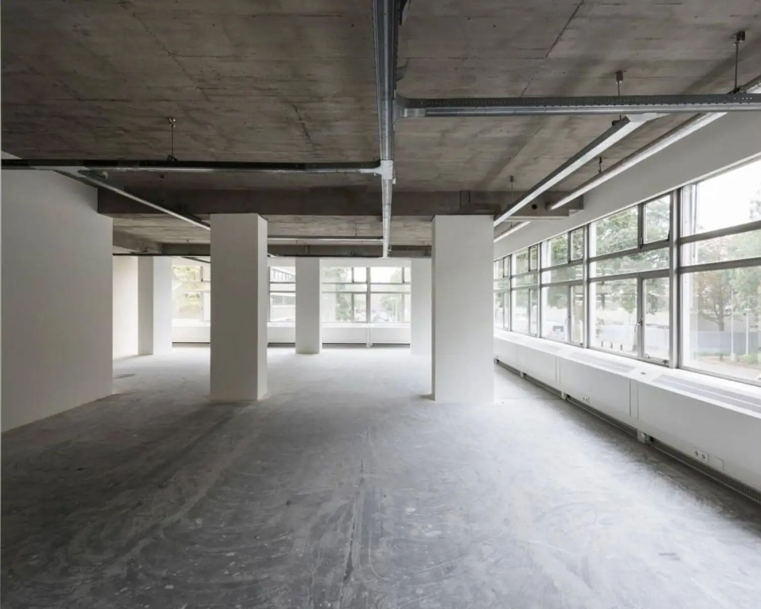 Lege kantoorruimte met betonnen plafond, steunpilaren en grote ramen langs de zijkant.