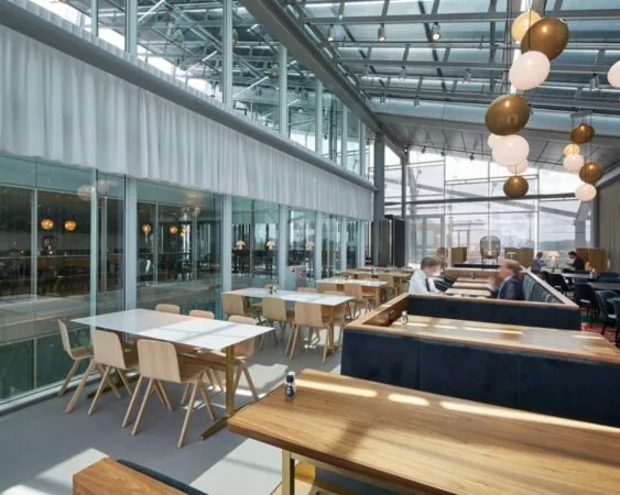 Moderne, lichtovergoten bedrijfsrestaurant met houten tafels, glazen wanden en hangende designlampen.