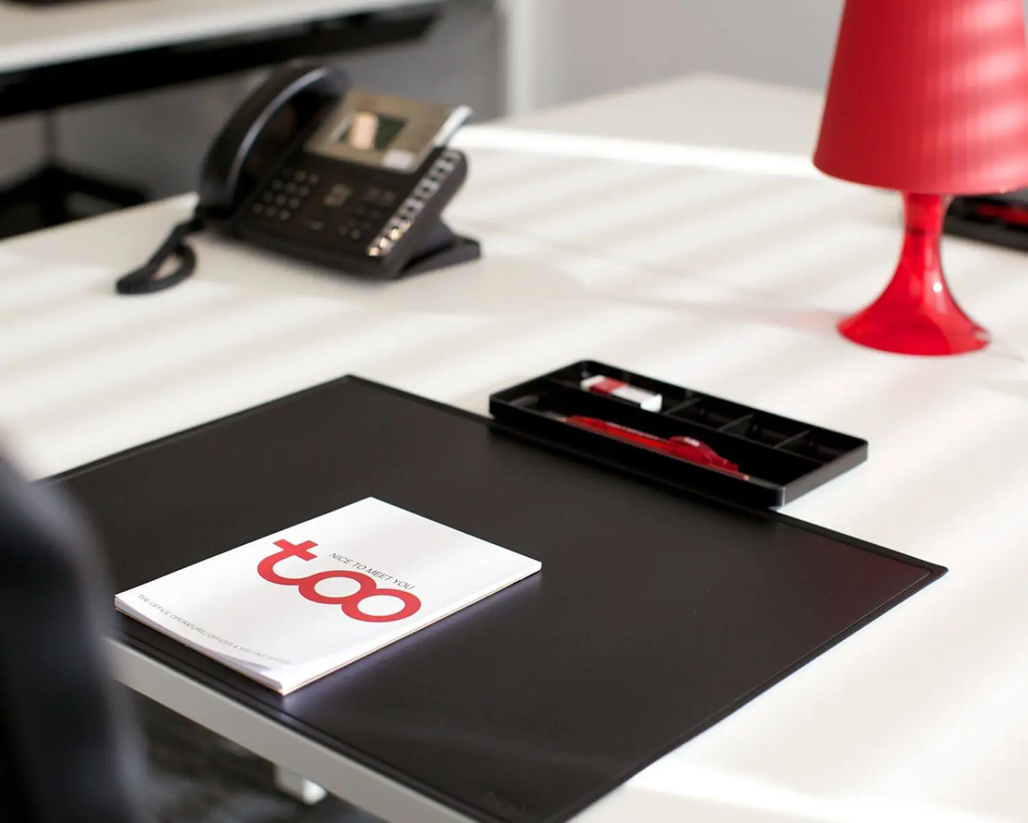 Modern bureau met zwarte bureaumat, rode bureaulamp, telefoon en een brochure met de tekst "too – nice to meet you".