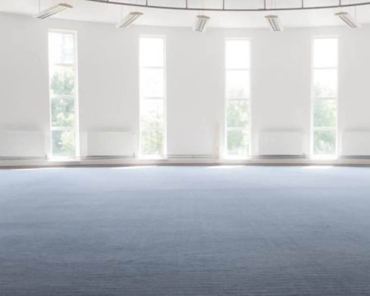 Lichte, lege ruimte met grote ramen, witte muren en een blauwe vloerbedekking.