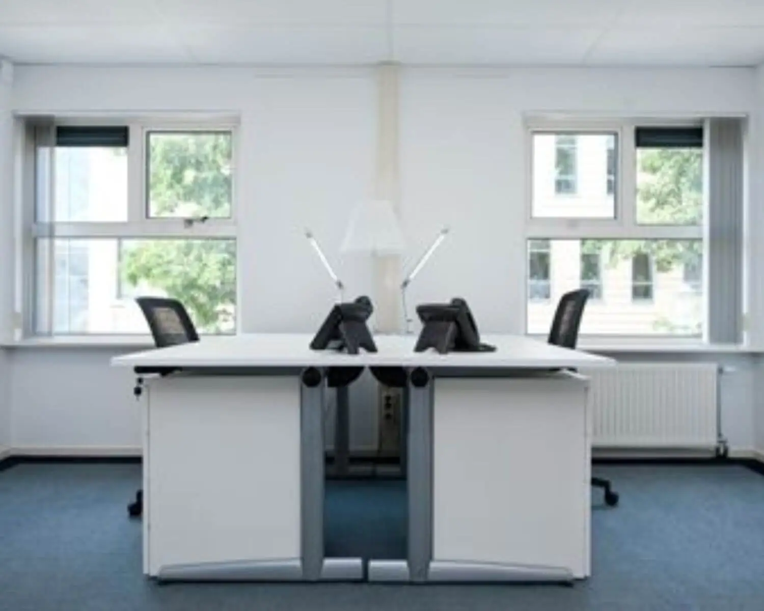 Twee tegenover elkaar staande bureaus met telefoons, bureaustoelen en bureaulampen in een helder verlichte kantoorruimte.