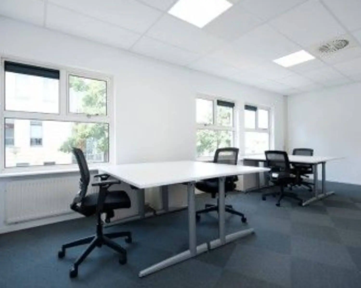 Lichte, moderne kantoorkamer met vier bureaus en bureaustoelen bij grote ramen.