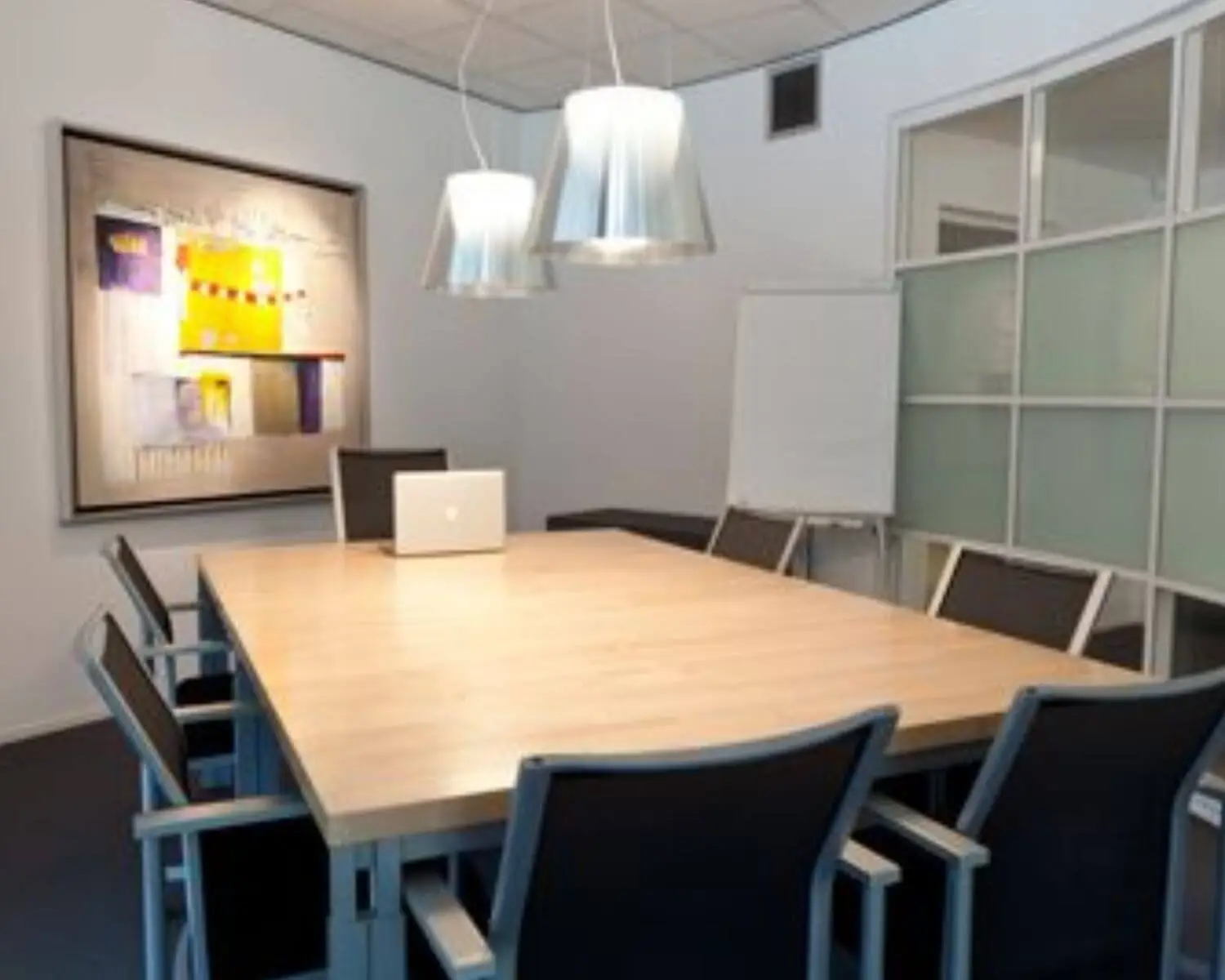 Vergaderruimte met houten tafel, moderne stoelen, hanglampen, een laptop, flip-over en abstract schilderij aan de muur.