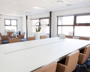Moderne vergaderruimte met een groot wit vergadertafel, bruine stoelen en een open zithoek op de achtergrond.