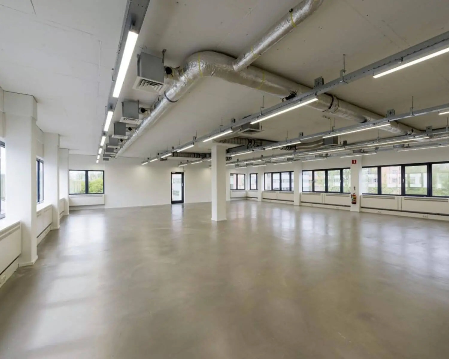 Lege, moderne kantoorruimte met betonnen vloer, witte muren en zichtbare ventilatiebuizen aan het plafond.
