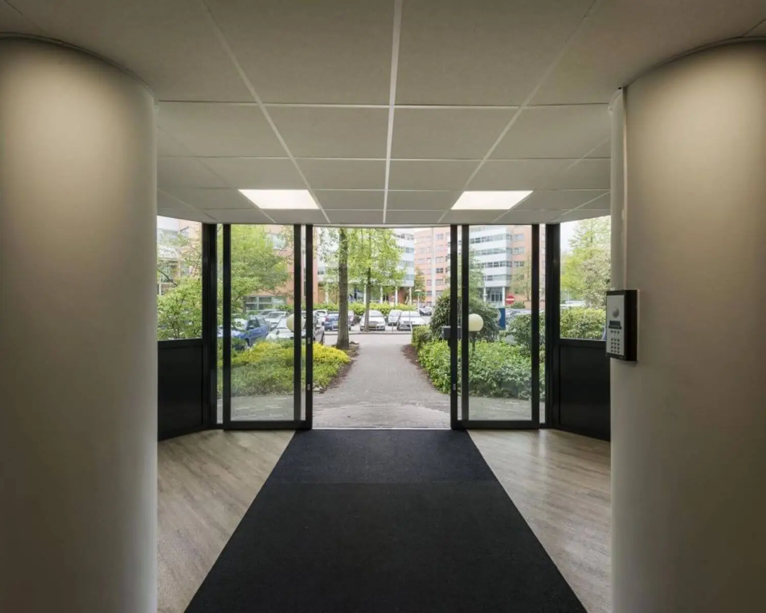 Ingang van een modern kantoorpand met glazen schuifdeuren en uitzicht op een parkeerterrein met groen.