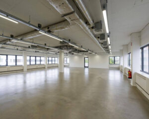 Lege, moderne kantoorruimte met grote ramen, betonnen vloer en zichtbaar plafond met ventilatiebuizen.