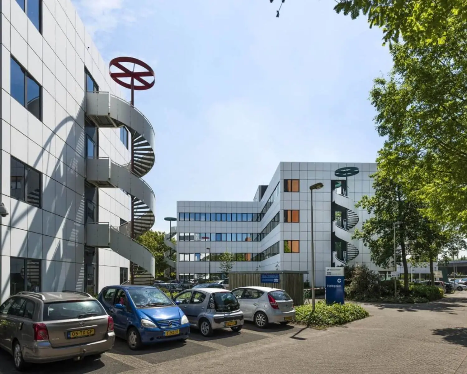 Kantoorgebouwen met spiraalvormige noodtrappen en geparkeerde auto's op een zonovergoten parkeerterrein.