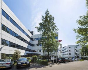 Modern kantoorgebouw met witte gevel en spiegelramen, omringd door geparkeerde auto's en bomen op een zonnige dag.